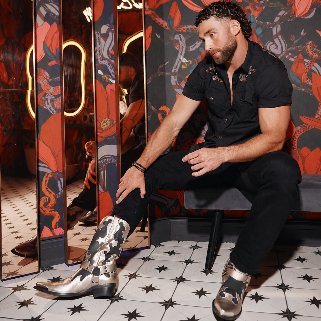 A man wearing a black short-sleeve button-up shirt, black pants, and silver cowboy boots with a black and white camo pattern sits on a bench in front of a red and black floral patterned wall with mirrored panels.